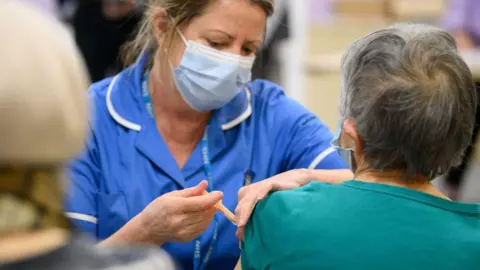 Getty Images The image shows a healthcare setting where a medical professional is administering an injection to another person’s upper arm. The medical professional is wearing a blue uniform, a face mask, and holding a syringe carefully while preparing or delivering the shot. The seated individual has their sleeve slightly pulled up to expose the upper arm for the injection. The background appears softly blurred, with other people and elements suggesting a busy clinic or vaccination site.