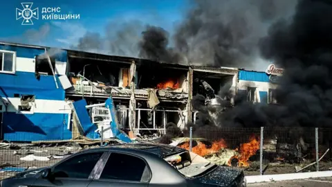 EPA/Shutterstock Black smoke is seen rising from a building, where some of the rooms are exposed and flames can be seen inside. A car is on fire in front and another car is seen in the foreground. 