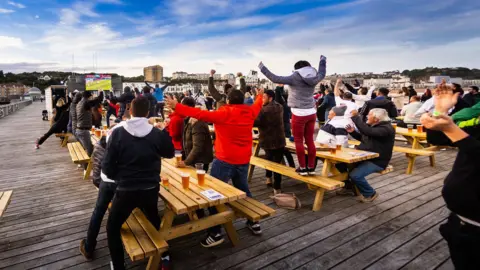 4TheFans People celebrating on Hastings Pier with screen at far end showing football match 