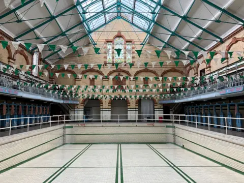 Inside empty Victorian building the Victoria Baths. The pool area is tiled with green and white and tiles and there's a white railing that goes all the way around. Green and white bunting hangs above and there is a partial glass roof.