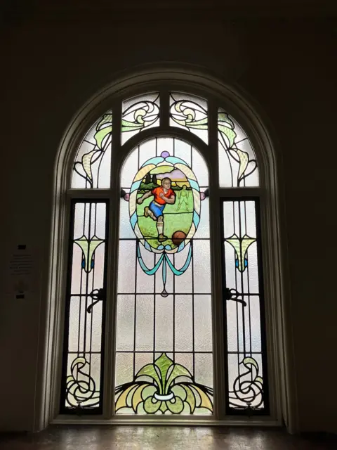 A close-up of a stained window with a middle panel showing a footballer about to kick a ball.