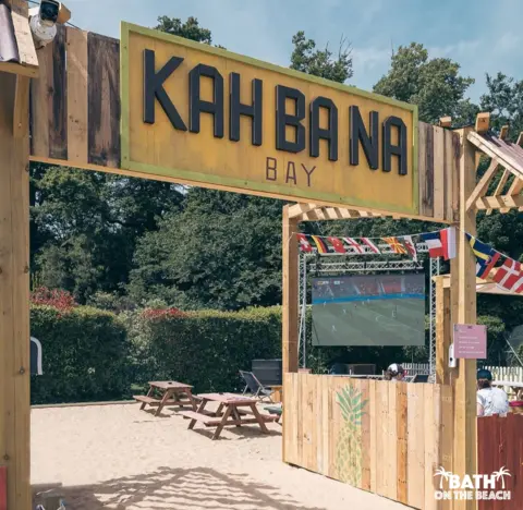 Bath on the Beach An outdoor beach-style bar with sand, benches and a screen showing the football