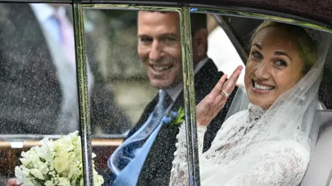 Reuters A woman is wearing a bridal dress sat in a car, smiling, and is sat next to a man in a wedding suit.