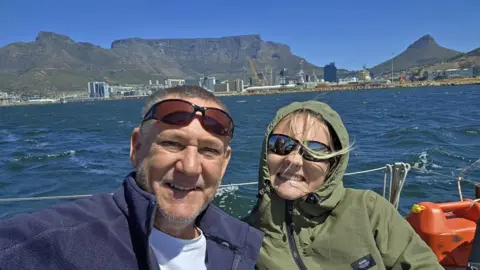 Rob Donald Man with moustache and trimmed beard wearing sunglasses on his brow smiling at the camera. He is wearing a fleece and a white t-shirt. Sitting next to him is a girl with her hair flying in the wind. She has a hood up on a waterproof she is wearing. She is smiling and they are both on a yacht. Behind them is Table Mountain.