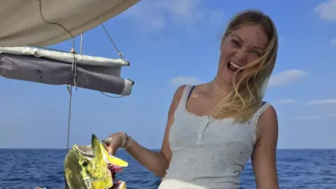 Rob Donald A girl is seen holding a vibrant fish by the gills she is has a big laugh. The background shows the boat's sail and the ocean. 