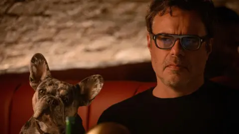 Disney Jemaine Clement as Steve sitting on a sofa with a dog next to him

