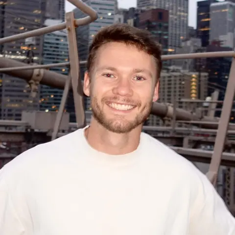 Daniel Bayen A man with short blonde hair and a beard, wearing a white t shirt and smiling at the camera. He is stood on a bridge with city skyscrapers behind him.