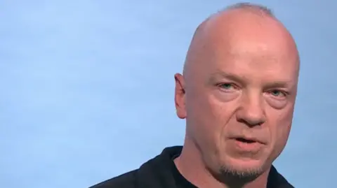 Fabulous A bald man with stubble, wearing a black shirt and looking at the camera with a serious expression. He is against a plain pale blue background. 