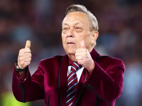 Getty Images Man in a claret-coloured blazer, and claret and blue tie, stands with both his thumbs pointing up. The background is blurred. 