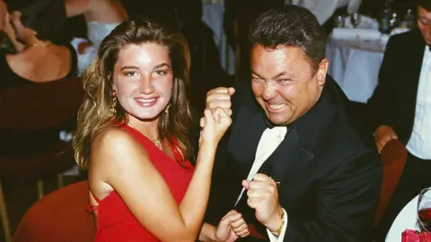 Getty Images Karren Brady, wearing a red dress and and David Sullivan, wearing a black suit and bow tie, pose for a picture at a function in 1993.