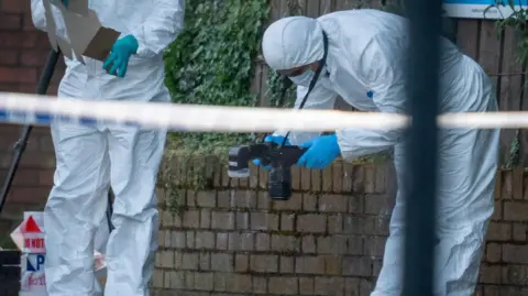 Aodhán Roberts Men in white forensics suits inspect crime scene. One is bent over at the hips taking a photo of evidence. 