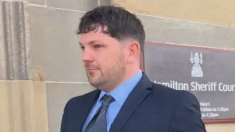 Tom Nicholson Jr has short, brown hair and is wearing a dark navy suit with a blue shirt and grey tie outside Hamilton Sheriff Court