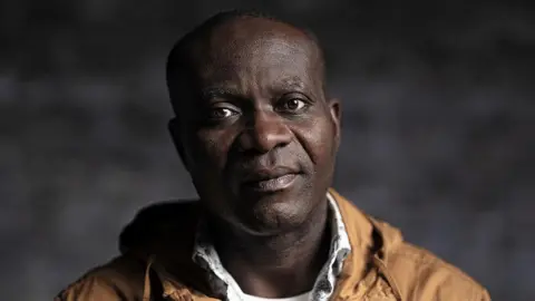 Gavin Hopkins Augustus Mensah is bald and looks at the camera with a neutral expression. He is wearing a brown jacket and white shirt underneath