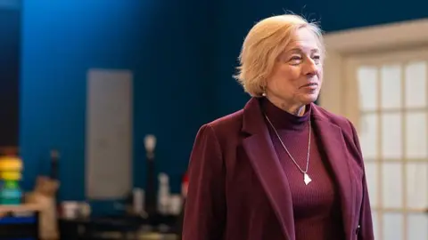 Getty Images Janet Mills, in a burgundy ensemble, looks off into the distance. She is wearing a silver necklace around her neck with a pendant in the shape of Maine. 