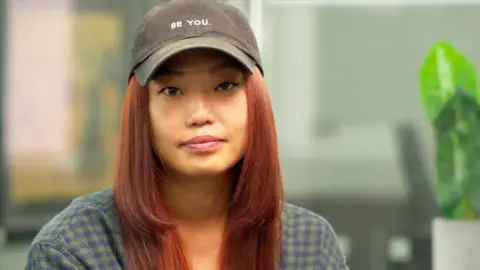 Jennifer An pictured looking at the camera, wearing a green and blue shirt and a baseball cap with the slogan "Be You"