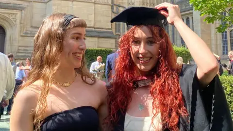 PA Media Grace and Caitlin stand in front of a university building together. Grace has long blonde hair and wears a black dress. Caitlin has died red hair and is wearing a graduation hat and gown. 