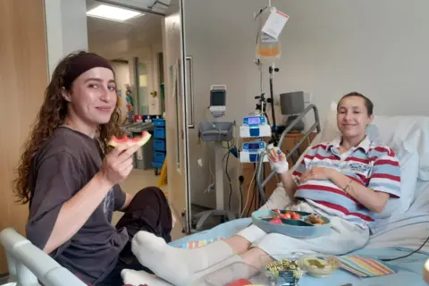 Collect/PA Real Life Caitlin and Grace Leggett sit on a hospital bed, smiling and eating fruit. Caitlin is laying beside medical equipment and an IV pole.