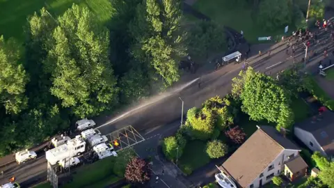 An aerial view of the water cannon in action. It is pushing back people who have gathered on the road. There are trees and houses on either side of the road.