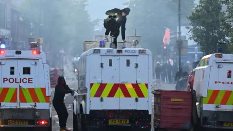 Getty Images Three police vans in a row across a road. Two people, dressed in black, are beside the middle van - one of them is on top of it. The background is smoke-filled and there are people to the right of the picture.