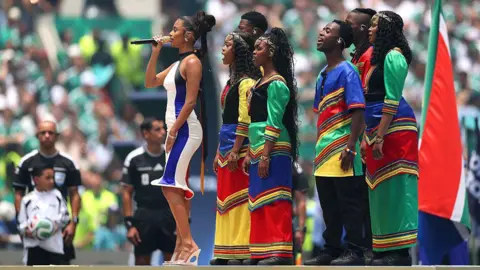 Getty Images Tyla, in a blue and white stripped dress, sings into a microphone in front of performers in green, red, blue and yellow outfits. 