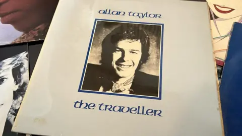 Allan Taylor's LP The Traveller showing a blue cover with a black & white photo of him smiling wearing a neckercheif in a frame in the centre