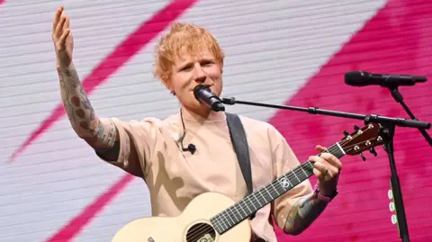 Getty Images Ed Sheeran singing into a microphone on stage with one hand on the neck of his guitar and the other raised.