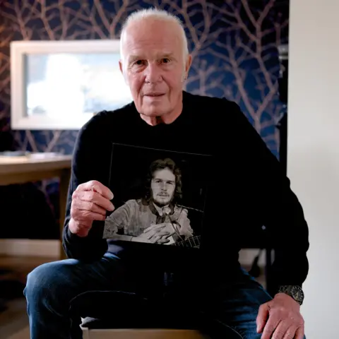Allan Taylor seated and looking into the camera, holding up a black & white photo of himself in the 1970s
