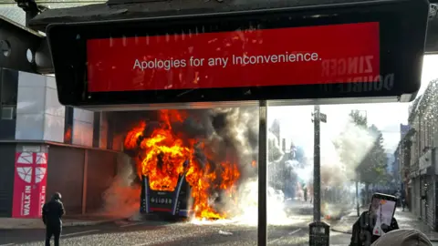 PA Media A Glider bus is on fire on a street. There is a sign at a bus stop at the top of the picture which says - Apologies for any Inconvenience. At the bottom right hand corner, some is filming the bus on a mobile phone.