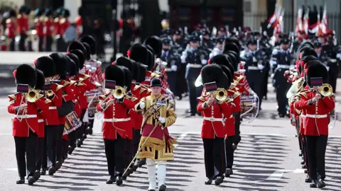 Getty Images Musicians from the Massed Bands of the Household Division parade during Trooping The Colour on June 13, 2026 in London, England. Four lines of musicians walk wearing a full red, white and black uniform, all of which are playing brass instruments. They are led by a man in the front and centre, wearing a smaller hat and wearing a sash.