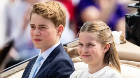 Getty Images Princess Charlotte and Prince George ride in a carriage with a tan interior. She is wearing a white dress with a white bow in her hair and is smiling. He is wearing a dark suit with a white shirt and a light blue tie.