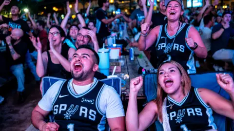 Brandon Bell/Getty Images People sitting outdoors face the camera wearing Spurs jerseys looking animated and smiling while cheering