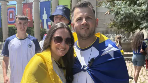 Husband and wife Admir and Alisa Maric wearing blue and yellow flags as capes as they stand outside a football stadium