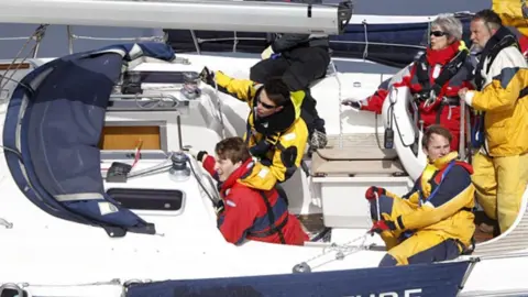 Jane and Alan Kelvey Five people can be seen sailing a boat, wearing red and yellow foul weather gear 