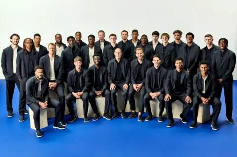 Marks and Spencer England men's squad wearing dark jackets and trousers and dark loafers. Some are seated in the front row while the rest of the team stand behind them