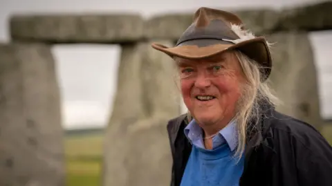 Tony Jolliffe/BBC News Archaeologist Phil Harding, who used to appear on Channel 4's "Time Team" programme, wearing his trademark hat with two feathers pushed through the headband on his left hand side. His long fair hair is hanging down on each side of his very characterful face. He is wearing a blue shirt and sweatshirt, with a coat that is open over the top. Behind him, out of focus, the outer circle of Stonehenge is visible and beyond that the grass of Salisbury plain. 