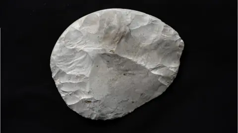 Tony Jolliffe/BBC News A rounded piece of flint that has been carefully worked to create a knife. It is white, with some brown and grey marks, and is set against a black background. The areas where pieces of the flint have been struck off using another stone are clearly visible. It is not a perfect circle but its sides are rounded. 