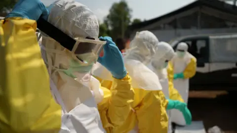 Amensisa Ifa / BBC A man in yellow and white PPE, including eye protection and blue gloves. Three other people in similar gear can be seen behind him - Bunia.