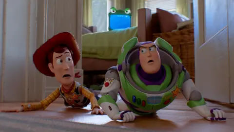 Disney Woody and Buzz Lightyear in Toy Story 5, looking scared and crouching on the ground, with green tablet Lilypad seen in the background on the bed