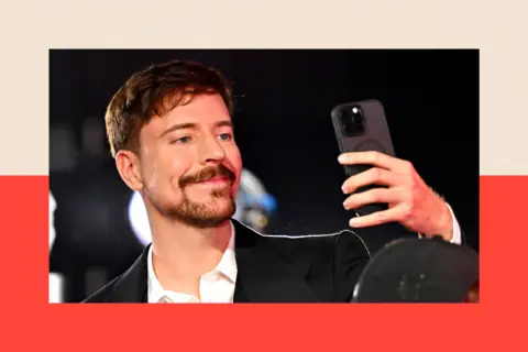 Getty Images YouTube star MrBeast attends the 2025 MTV Video Music Awards. He is a white man with dark hair and a dark beard. He is wearing a dark suit jacket and a white open-necked shirt and is holiding a mobile phone in front of him as if taking a selfie.