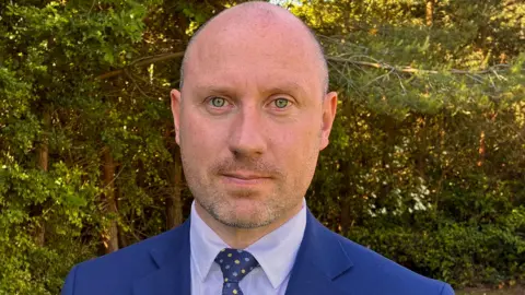 A balding man with a stubble and wearing a blue suit looking directly at the camera with trees in the background