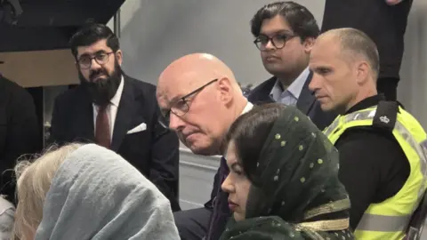 PA Media Group of people seated closely together in an indoor meeting setting, including individuals in formal attire and one person in a high-visibility police vest, engaged in discussion while others listen attentively around them.