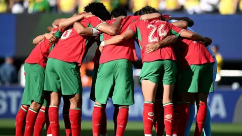 Getty Images Morocco players in red shirts and green shorts form a huddle before their 2026  World Cup match against Brazil in New Jersey on 13 June.