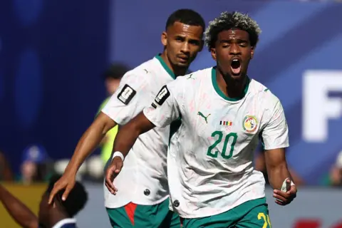 FRANCK FIFE / AFP via Getty Images Ibrahim Mbaye celebrates after scoring for Senegal against France in the 2026 World Cup on 16 June. He is wearing a white top and dark green shorts, and his mouth is open as he runs towards the camera.
