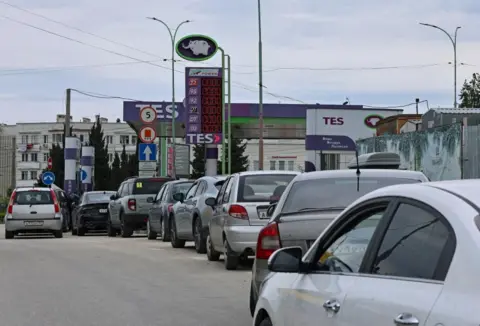 Reuters A line of silver and grey cars snaking round from a petrol station where the prices electronic display has no numbers on it.