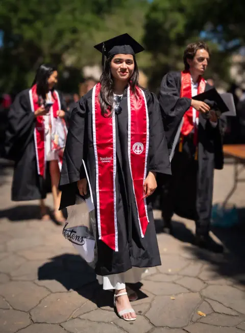 This is a full length photo of Ifdita Hasan who is dressed in a graduation gown and wearing a mortar board on her head. There are two more students similarly dressed behind her.