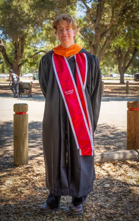 This is a full length photo of Lucy Zimmerman who is dressed in a graduation gown. There are trees behind her.
