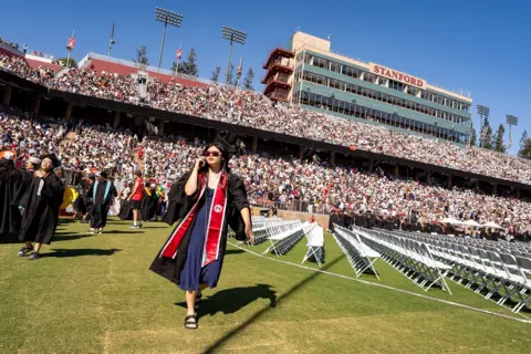A woman in a graduation gown is walking on the grass in front of a huge stand filled with people. She has a cellphone against her ear and appears to be speaking on it. 