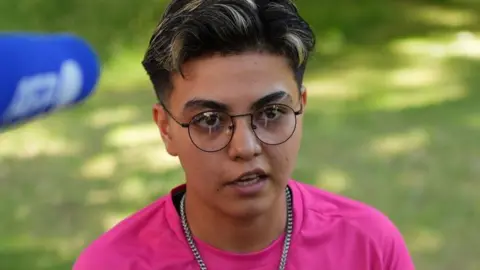 Reuters Ekil Latifi, a woman with cropped dark hair with highlights, wearing black wire-framed glasses and a silver chain necklace with a bright pink T-shirt 