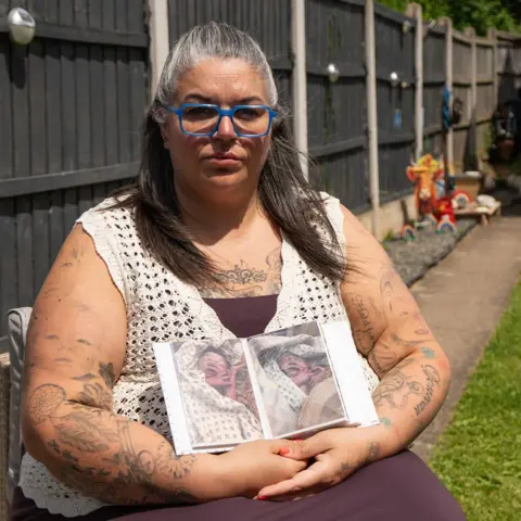 BBC/Chris Waring Natalie's Needham, who wears blue glasses, sits in her garden and holds two pictures of Kouper. A fence can been seen behind her.