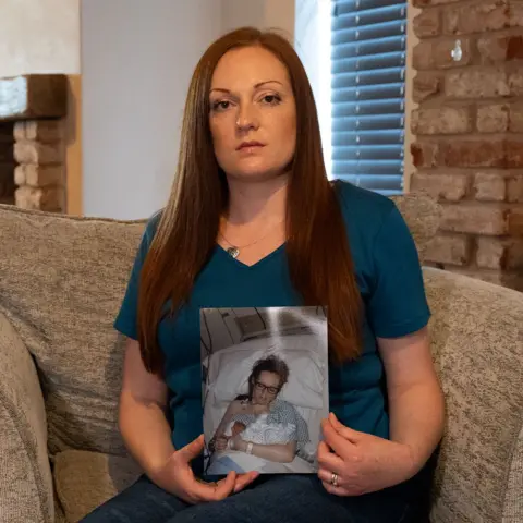 BBC/Chris Waring Felicity Benyon sits in an armchair holding a picture.